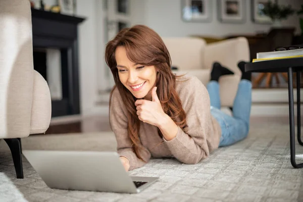 全长拍摄快乐女人在家里放松的时候使用笔记本电脑和在网上浏览 穿着毛衣和蓝色牛仔裤的迷人女人 — 图库照片