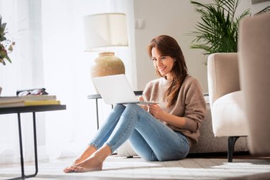 Orta yaşlı çekici bir kadının dizüstü bilgisayar kullanırken yerde oturması. Güzel bir kadın günlük kıyafetler giyiyor ve video görüşmesi yapıyor. Merkez Ofis.