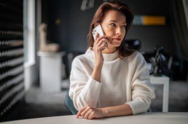 Orta yaşlı iş kadını ofiste masa başında otururken telefonla konuşuyor. Yönetici iş kadını süveter giyiyor.. 