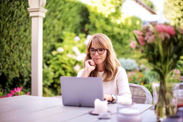 微笑的中年妇女坐在家里的阳台上 一边用笔记本电脑一边打电话 — 图库照片