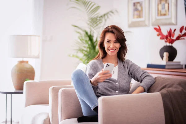 迷人的黑发女人穿着休闲装 在家里放松一下 并使用智能手机 微笑女性短信 — 图库照片
