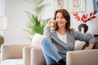 Evde dinlenirken ve akıllı telefon kullanırken gündelik kıyafetler giyen çekici esmer kadın. Gülümseyen bir kadın arıyor..