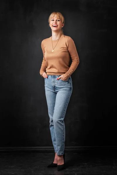 孤立した暗い背景に立つ魅力的なブロンドの髪の女性の完全な長さ 幸せな女性のセーターやジーンズを着て カメラと笑顔を見て スペースのコピー — ストック写真