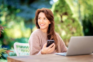 Confident middle aged woman text messaging and using laptop while sitting on balcony. Attractive female wearing sweater and cheerful smiling.