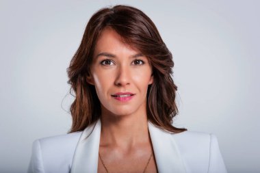 Headshot of an attractive middle aged smiling and looking at camera. Brunette haired female wearing white blazer while sitting at isolated dark background. Copy space. Studio shot.