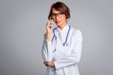 Portrait of smiling female doctor using mobile phone and talking. Isolated grey background. Confident healthcare worker is wearing lab coat. 