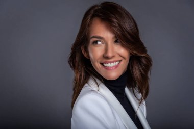 Headshot of an attractive middle aged woman with toothy smile wearing white blazer while standing at isolated dark background. Woman looking away. Copy space. Studio shot.