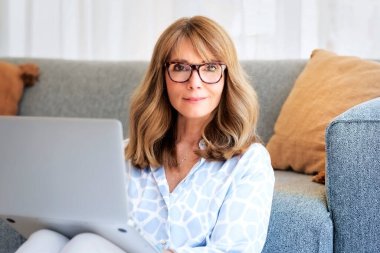 Orta yaşlı bir kadının iş için dizüstü bilgisayarını kullanırken yakın plan fotoğrafı. Evde çalışırken günlük kıyafetler ve gözlük takan sarı saçlı iş kadını. Merkez Ofis. 