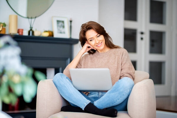 Shot of an attractive middle aged woman sitting in an armchair while using laptop and having video call. Beautiful female wearing casual clothes.
