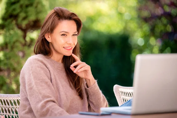 一位自信的中年妇女坐在家里的阳台上 一边用笔记本电脑 迷人的女人 穿着毛衣 开朗地微笑着 — 图库照片
