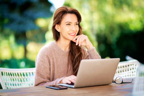 自宅のバルコニーに座っている間 ラップトップを使用している中年の女性を自信を持ってください 魅力的な女性はセーターを着て陽気な笑顔 — ストック写真