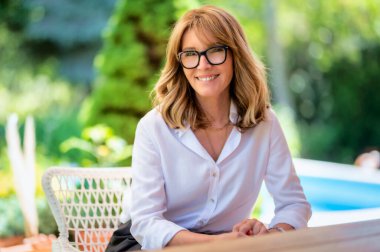 Close-up portrait shot of cheerful middle aged woman wearing glasses and white shirt while relaxing on the patio at home.