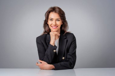  İzole edilmiş gri arka planda otururken siyah ceket giyen bir iş kadını. Uzayı kopyala Eller çenede..