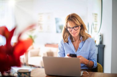 Middle aged woman sitting at her dining room table and using earphone and laptop while having video call. Attractive female holding smartphone in her hand. Home office. 