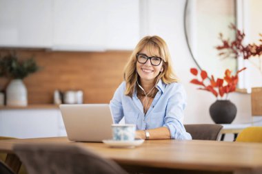 Orta yaşlı bir kadın yemek masasında oturmuş video görüşmesi yaparken kulaklık ve dizüstü bilgisayar kullanıyor. Merkez Ofis. 