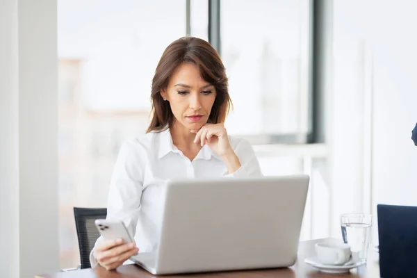 考虑用智能手机和笔记本电脑工作的女商人 微笑着坐在办公室和工作的女性 专业女性看上去很体贴 — 图库照片