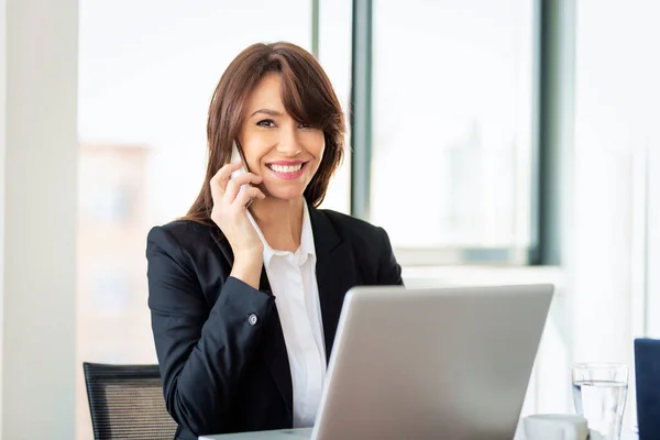 有吸引力的女商人有一个电话和使用笔记本电脑的工作 微笑着坐在办公室和工作的女性 穿着运动衫和白衬衫的行政专业女性 — 图库照片