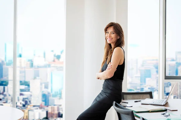 役員室のオフィスデスクに立つ魅力的なビジネスウーマン 黒のドレスと明るい笑顔を身に着けているエグゼクティブ女性 — ストック写真