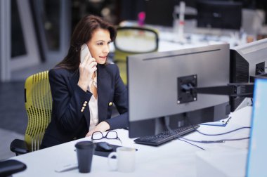 Orta yaşlı iş kadını bilgisayar kullanıyor ve ofiste masa başında otururken telefonla konuşuyor. Profesyonel kadın yeni iş projesinde çalışıyor..