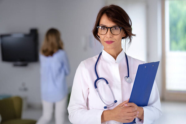 Female doctor wearing lab coat and stethoscope and holding clipboard in her hands  while standing at hospital corridor