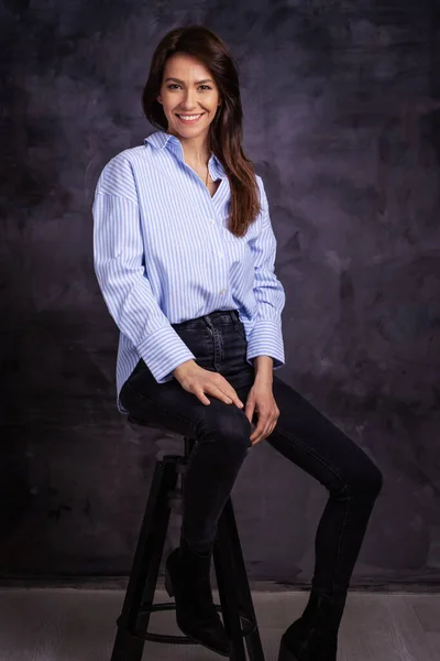 Studyjne Ujęcie Atrakcyjnej Kobiety Koszuli Dżinsach Siedzącej Odosobnionym Ciemnym Tle — Zdjęcie stockowe