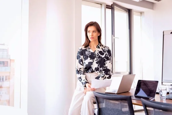 ビジネスカジュアルを着てモダンなオフィスのデスクに座っている思考ビジネス女性のショット プロの女性は思慮深く見て 彼女の手で文書を保持 — ストック写真