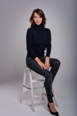 Gri arka planda iskelede otururken boğazlı kazak ve siyah pantolon giyen çekici esmer kadının uzun pozu. Boşluğu kopyala.