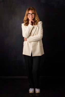 Çekici, orta yaşlı, gülümseyen ve kameraya bakan bir kadın. Sarışın kadın, izole edilmiş koyu arka planda dikilirken ceket ve pantolon giyiyor. Uzayı kopyala Stüdyo çekimi. 