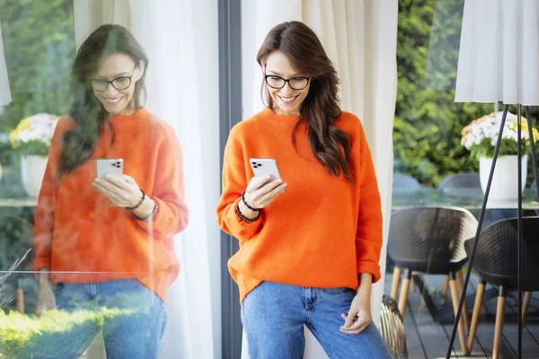 站在家里的窗口时 照片上有一个快乐的女人在发短信 穿着橙色毛衣和蓝色牛仔裤的布鲁内特妇女 — 图库照片