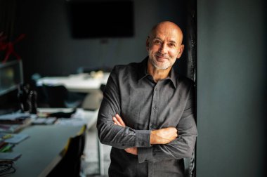 Ofiste kollarını kavuşturmuş duran orta yaşlı iş adamının portresi. Kendine güvenen profesyonel bir erkek tişört giyiyor ve kameraya bakıyor..