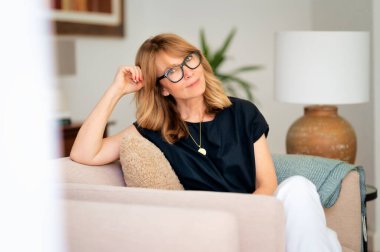 Çekici, orta yaşlı bir kadının evdeki koltukta dinlenirken yakın plan portresi. Sarı saçlı, gözlüklü ve siyah gömlekli bir kadın..