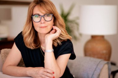 Çekici, orta yaşlı bir kadının evdeki koltukta dinlenirken yakın plan portresi. Sarı saçlı, gözlüklü ve siyah gömlekli bir kadın..