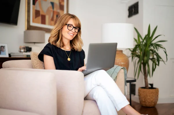 有魅力的中年妇女坐在家里用笔记本电脑的肖像 金发女人戴着眼镜和黑色衬衫 总部办公室 — 图库照片
