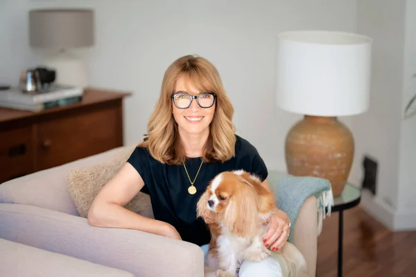 有魅力的中年女性在家里的扶手椅上放松的画像 金发女性 戴着眼镜 穿着黑色衬衫 她的小狗坐在她的腿上 — 图库照片