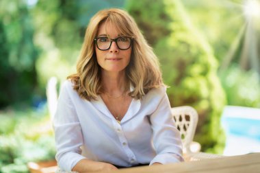 Close-up portrait shot of cheerful middle aged woman wearing glasses and white shirt while relaxing on the patio at home.
