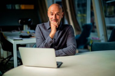 Orta yaşlı bir işadamı modern bir ofiste oturuyor ve dizüstü bilgisayarını iş için kullanıyor. Tişört giyen profesyonel bir adam..