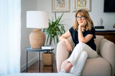 Çekici orta yaşlı bir kadının yakın plan portresi. Evdeki koltukta dinleniyor ve derin düşüncelere dalmış. Sarı saçlı, gözlüklü ve siyah gömlekli bir kadın..