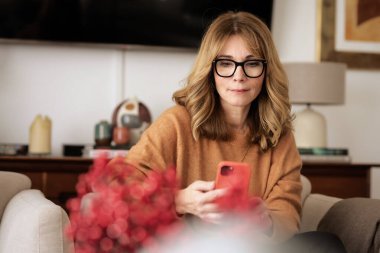 Cep telefonu ve kısa mesaj kullanan orta yaşlı bir kadının yakın çekimi. Kendine güvenen bir kadın modern bir evde koltukta dinleniyor..