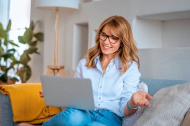 Sarışın, orta yaşlı bir kadın evdeki koltukta oturuyor ve dizüstü bilgisayar kullanıyor. Kendine güvenen bir kadın video görüşmesi yapıyor.. 