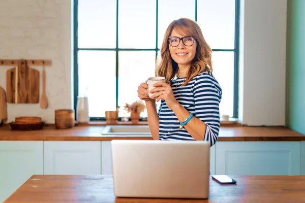 台所で朝食を準備しながら ラップトップを使用してビデオ通話をしている中年の女性のショット 内務省だ 幸せな女性保持Aマグカップで彼女の手 — ストック写真