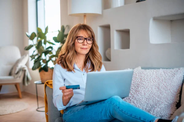 中年妇女坐在家里 用信用卡和笔记本电脑上网购物 自信的女性 身穿衬衫和眼镜 — 图库照片