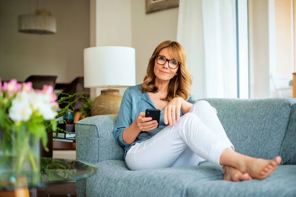 一个有魅力的中年女人在家里放松一下 发短信 全身而退 自信的女人坐在沙发上 穿着休闲装 — 图库照片