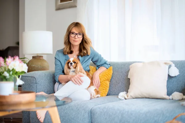 一个快乐的中年妇女带着她可爱的小狗在家里的沙发上放松 穿着休闲装和眼镜的自信女性 — 图库照片