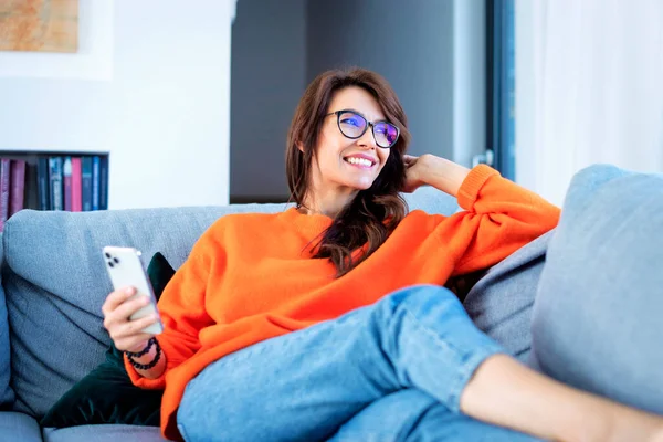 拍了一个迷人而快乐的女人使用她的智能手机和短信 同时放松在沙发上的家 穿着橙色毛衣的漂亮女人 — 图库照片