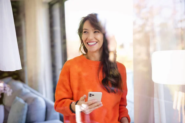 自宅の窓の前に立ちながら朗らかな女のテキストメッセージのポートレートショット 自信に満ちた女性はオレンジのセーターと明るい笑顔を着て — ストック写真