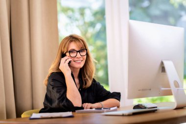 Orta yaşlı bir kadının masada oturup cep telefonu kullanması. Kendine güvenen, gözlük takan, günlük kıyafetler giyen ve evden çalışan bir kadın. Merkez Ofis.