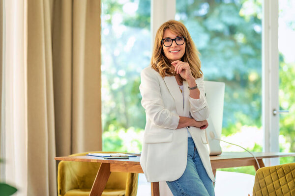 Portrait of mid aged businesswoman standing at desk while looking at camera and smiling. Confident female wearing blazer and glasses. Home office. 