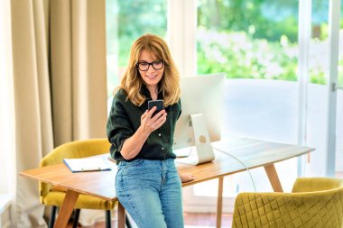 Orta yaşlı bir kadının masada durup cep telefonu kullanması. Gözlük takan, sıradan kıyafetler giyen ve mesajlaşan kendinden emin bir kadın. Merkez Ofis.