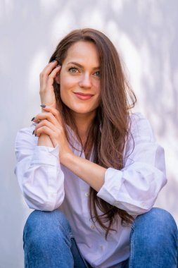 Güneşin altında oturmuş gülümseyen ve kameraya bakan güzel bir kadının portresi. Beyaz gömlek ve kot pantolon giyen çekici bir kadın..