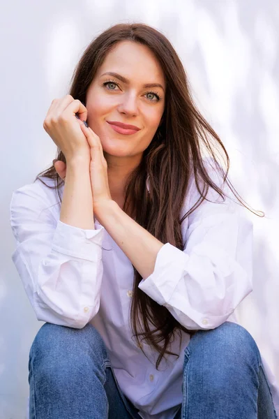 一个美丽的女人坐在阳光下 微笑着看着相机的画像 穿着白衬衫和蓝色牛仔裤的迷人女性 图库图片